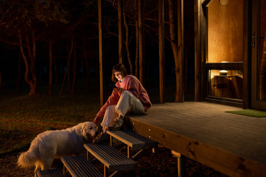 Young woman with dog sits on porch of a wooden house during a night time, enjoying nature while resting in cottage at countryside