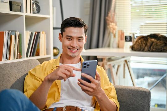 Handsome and happy millennial Asian man using his smartphone, relaxing on sofa in living room.