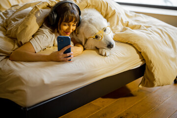 Obraz premium Young woman takes selfie photo or has a video call while lying with her cute dog under a blanket in bed. Spending leisure time and having fun together. Friendship with pets and coziness concept