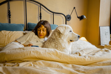 Obraz premium Young woman uses a smartphone while lying with her dog in bed at cozy bedroom in beige tones. Leisure time and relaxation with pets concept