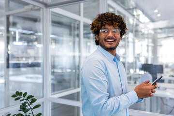 Portrait of successful hispanic programmer engineer developer inside office, man in shirt and glasses holding smartphone in hands smiling and looking out window, businessman at workplace young happy.