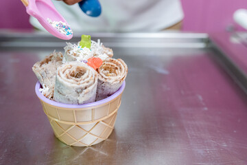 A worker is decorating a rolled ice cream tub with chips and teddy bear toppings with a spoon
