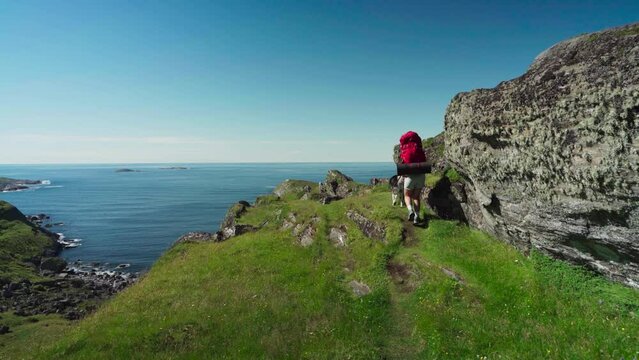 Traveler With Backpack And Alaskan Malamute Walking On The Rocky Mountains Near Lovund Island Village, Norway. Wide Shot