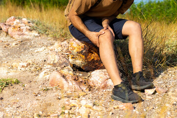 Caucasian man traveler suffering knee pain during hiking on mountain hill. Handsome guy with outdoor lifestyle walking on mountain dirt road on summer vacation. Travel health care insurance concept.