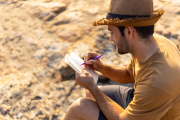 Caucasian man traveler writing journey memories in diary book on coastline hill at sunset. Handsome...