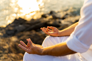 Caucasian man practicing meditation prayer with ocean nature on rocky coastal hill at summer sunset. Wellness people do outdoor self relaxing yoga exercise. Mental health care and motivation concept.