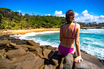 A beautiful girl in a pink bikini sits on a rock and enjoys the paradise scenery of small bays with golden sand and turquoise ocean in Sunshine Coast. Hidden gems in Queensland, Australia