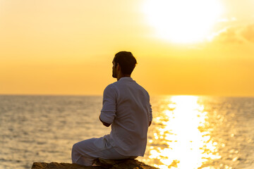 Caucasian man practicing meditation prayer with ocean nature on rocky coastal hill at summer sunset. Wellness people do outdoor self relaxing yoga exercise. Mental health care and motivation concept.