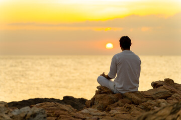 Caucasian man practicing meditation prayer with ocean nature on rocky coastal hill at summer sunset. Wellness people do outdoor self relaxing yoga exercise. Mental health care and motivation concept.