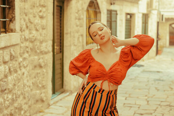 Beautiful stylish girl with long earrings, wearing an orange suit looks at the camera. Traveling around Europe Fashionable woman smiling in the old town and posing.