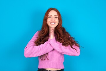 young woman wearing pink sweater over blue background keeps palms together, has pleased expression....
