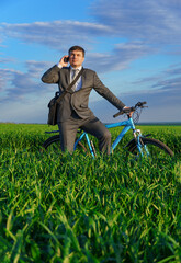 a businessman rides a bicycle on a green grassy field, dressed in a business suit, he makes a phone call, beautiful nature in spring, freelance business concept
