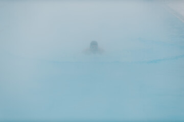 Fit swimmer male training swim in open winter swimming pool with fog. Geothermal outdoor spa health concept.