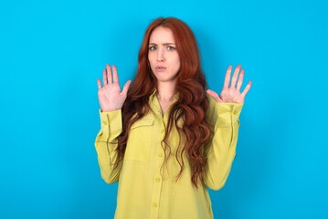 young woman wearing green sweater over blue background Moving away hands palms showing refusal and...