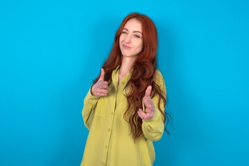 Joyful young woman wearing green sweater over blue background wink and points index fingers at...