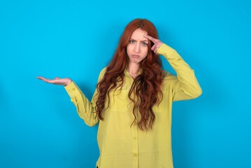 young woman wearing green sweater over blue background confused and annoyed with open palm showing copy space and pointing finger to forehead. Think about it.