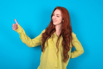 Fototapeta premium young woman wearing green sweater over blue background Looking proud, smiling doing thumbs up gesture to the side. Good job!