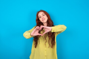 young woman wearing green sweater over blue background smiling in love doing heart symbol shape with hands. Romantic concept.