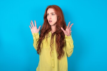 young woman wearing green sweater over blue background shouts loud, keeps eyes opened and hands tense.