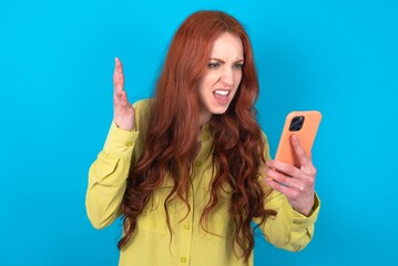Angry young woman wearing green sweater over blue background screaming on the phone, having an...