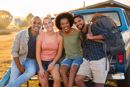 Portrait Of Friends Sitting On Tailgate Of Pick Up Truck On Road Trip To Cabin In Countryside