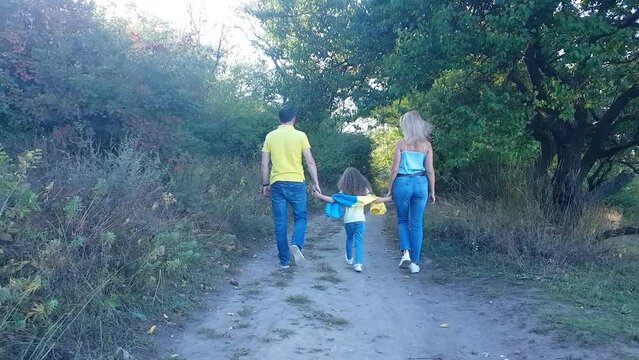 Family - father, mother and daughter go into the distance, holding hands, along the path in the park with the flag of Ukraine