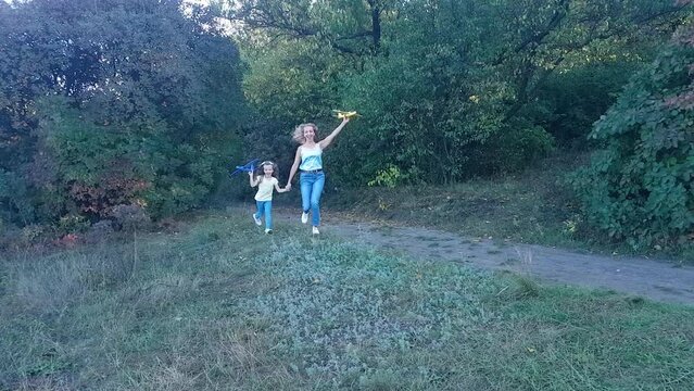 Happy Ukrainian mother and daughter run with yellow and blue toy airplanes in their hands in the park towards the camera and launch them