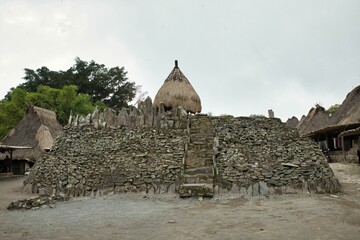 The traditional Bena Village on Flores, in the foreground a stone wall, behind it several cone-shaped thatched huts, in the background trees.