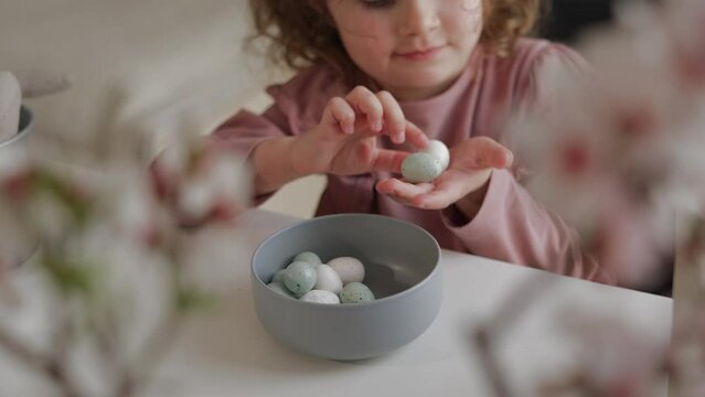 View of hands of pretty girl playing with Easter eggs. She is sitting at a table with spring flowers, almond blossom, looking and touching the eggs celebrating Happy Easter.