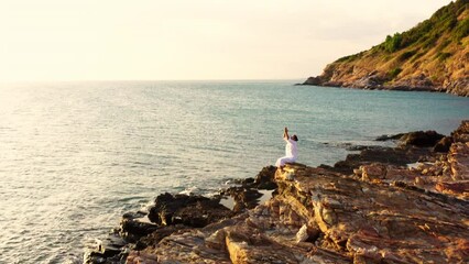 Aerial view of Caucasian man practicing meditation with ocean nature on rocky coastal hill at summer sunset. Wellness man do outdoor relaxing yoga exercise. Mental health care and motivation concept.