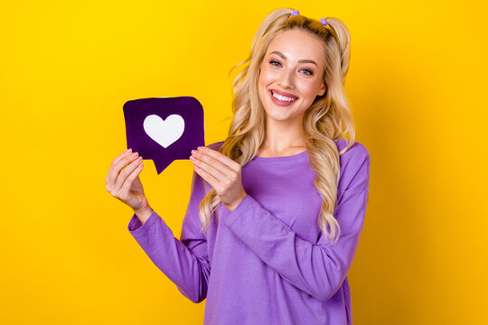 Photo portrait of pretty young girl hold heart icon notification symbol dressed stylish violet clothes isolated on yellow color background