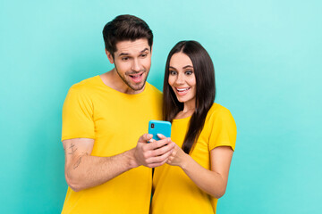 Photo of surprised lovely couple hold smartphone wear yellow t-shirts enjoy crazy black friday sale website isolated on aquamarine color background