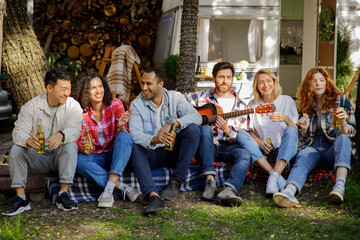 Handsome young man plays the guitar for friends. Mixed-race company of friends. Having fun listening to music at holiday summer parties near the travel trailer. Happy multi ethnic young people.