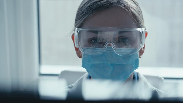 A female researcher sits at a workplace in a laboratory, behind a personal computer monitor. Against the background research statistics. Pharmaceutical medical worker in protection works at keyboard.