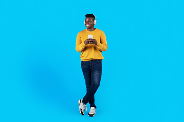 Fototapeta na wymiar Smiling Black Guy In Wireless Headphones Holding Smartphone And Looking Away