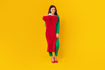 Full length shot of fashion designer woman standing with hanger in hand fitting on red elegant dress, yellow background