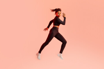 Fototapeta na wymiar Young fit black woman jumping, having cardio training in fitwear over peach neon background, full length, copy space