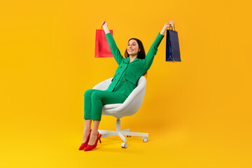 Full body length shot of contented shopaholic woman sitting on chair and raising hands with shopping bags, free space