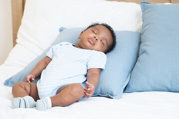 Cute African Nigerian newborn baby sleeping lying on pillow on white bed at home. Innocence infant with curly hair wear clothes and socks sleep in daytime nap