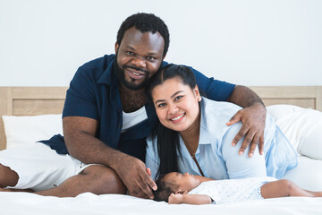 African father and Asian mother touching and lull cute newborn baby sleeping lying on bed at home, parent smiling looking at camera. Multiracial family bonding and child care concept