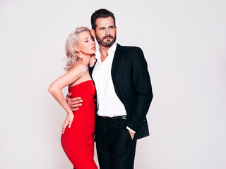 Beautiful fashion woman and her handsome boyfriend in black elegant suit. Sexy blond model in red evening dress. Fashionable couple posing in studio. Brutal man and his female. Isolated on white