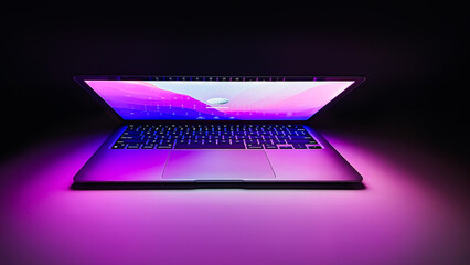 Laptop with half lid open on a table lit with colorful purple and blue desktop screen wallpaper in a dark room	
