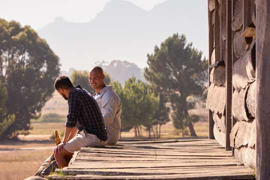 Two Male Friends On Vacation Sitting On Porch Of Countryside Cabin Drinking Beer Together