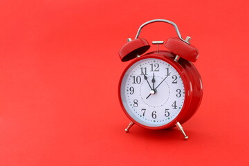 A red alarm clock stands on a red background. The time shows five minutes to twelve.	
