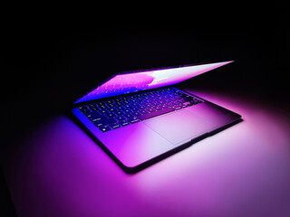Laptop with half lid open on a table lit with colorful purple and blue desktop screen wallpaper in...