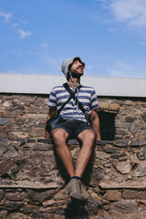 Happy young latin man sitting on a colonial stone wall looking at the sky. Vertical photo.