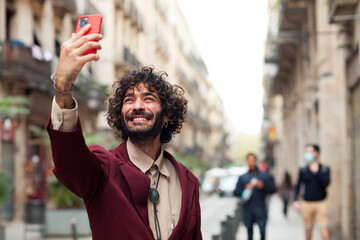 Handsome young man taking a self portrait while walking through the city. Urban millenial with beard and earring using a smart phone and smiling carefree outdoors. Copy space.