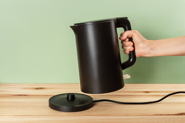 Matte black kettle kettle in a woman hand. Person hand taking off electric kettle from power base...