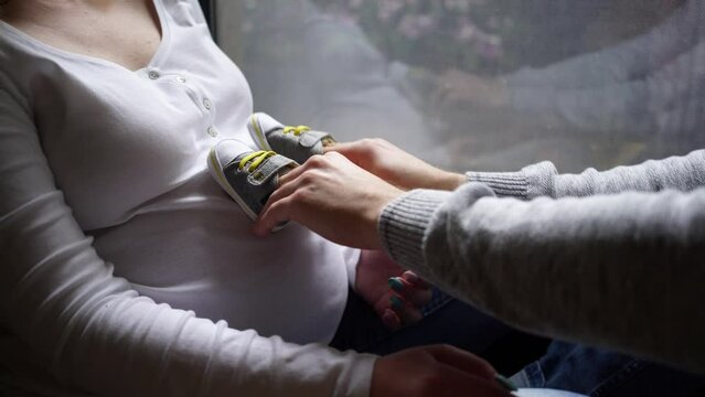 Unrecognizable pregnant woman with large tummy and husband in casual clothes sit on windowsill. Man holds small boots, walking on belly of pregnant wife, waiting for the newborn baby concept