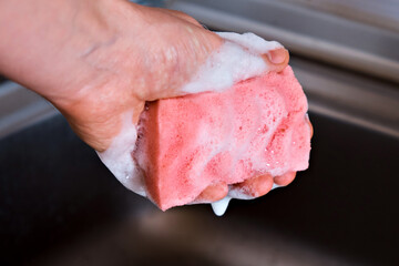 A man holds a red kitchen sponge in his hand, which is foamed from detergent, on the background of a stainless steel sink. Close-up